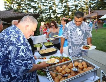 A crowd of people in bright, summery clothes gather around an outside buffet table
