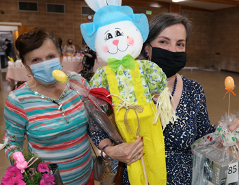 Two masked women hold up raffle prizes, including a bunny scarecrow. 