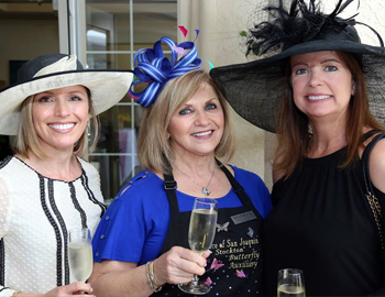 Three smiling women with fancy hats in white, blue and black. 