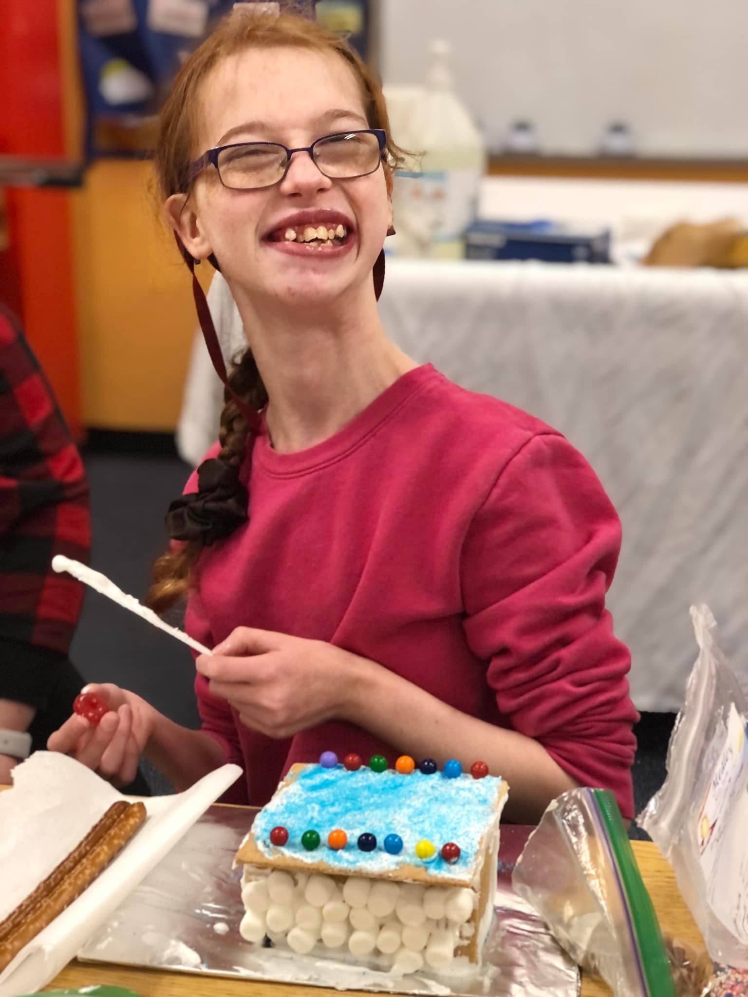 A teenage girl in a pink shirt and glasses smiles while building a gingerbread house.