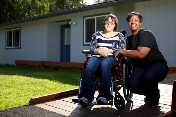 A woman in a wheelchair and her female support staff are on a wooden deck in front of a house on a sunny day