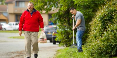A man in a t-shirt rakes the lawn, and another man in a red sweatshirt walks past him carrying a bag of groceries.