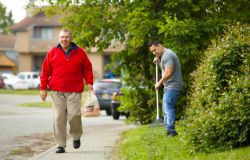 A man in a t-shirt rakes the lawn, and another man in a red sweatshirt walks past him carrying a bag of groceries.