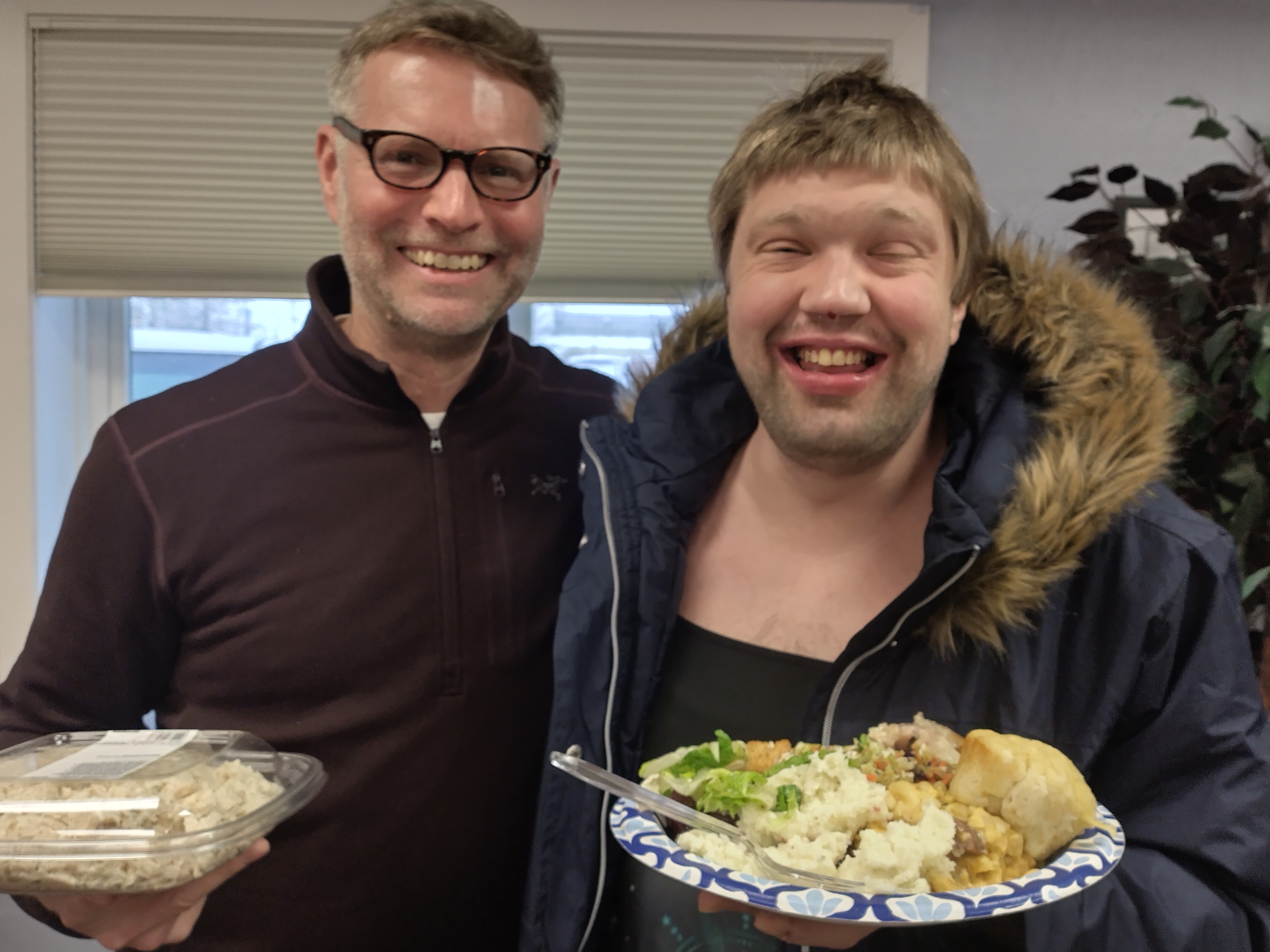 Two gentlemen smile and hold plates of Thanksgiving food