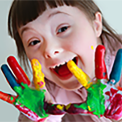 A young girl has red, yellow, green, and blue paint on her outstretched hands and a big smile on her face