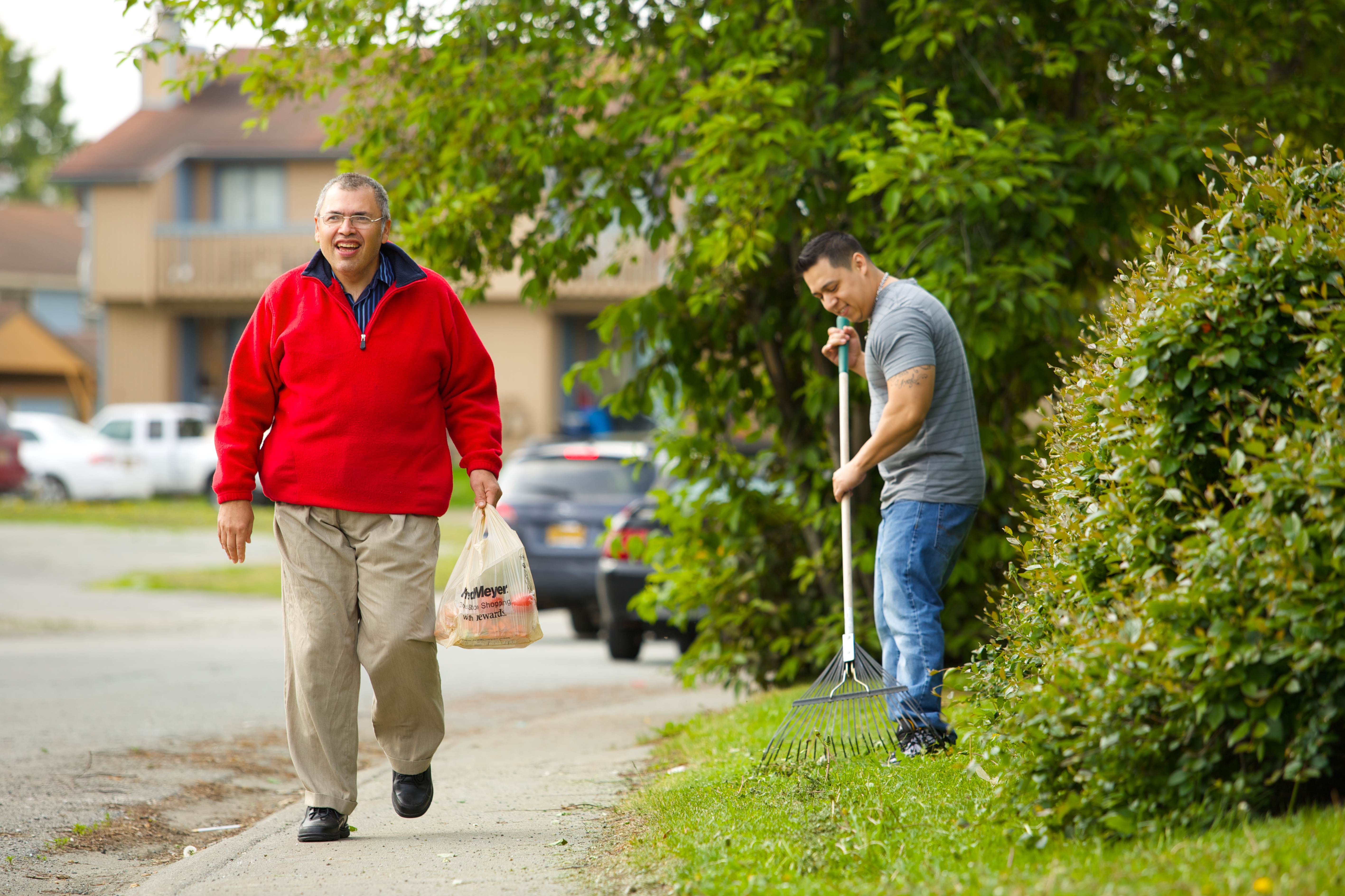 A man in a t-shirt rakes the lawn, and another man in a red sweatshirt walks past him carrying a bag of groceries.