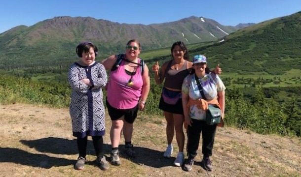 Four women stand together while hiking in Alaska. Behind them is a blue sky and green mountains.